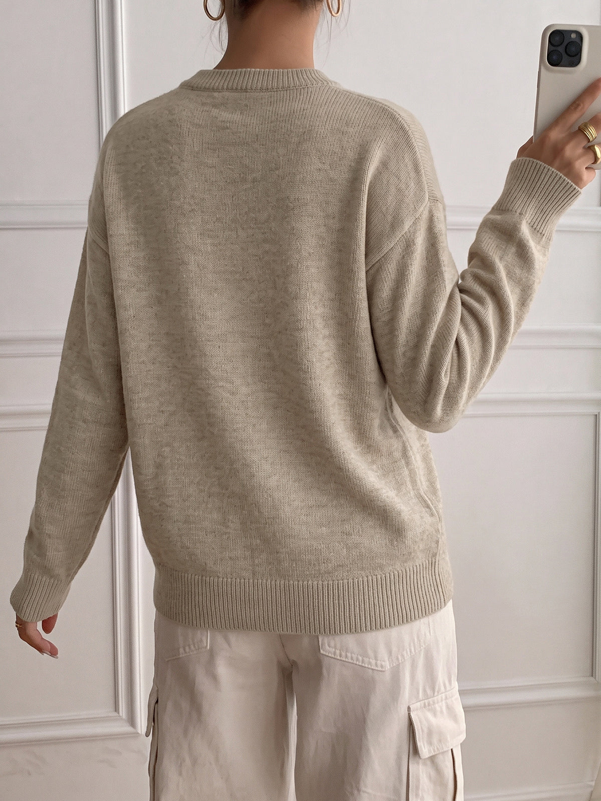 A person shown from the back wearing a light beige knit sweater and light cargo pants, holding a phone in front of a white paneled wall.