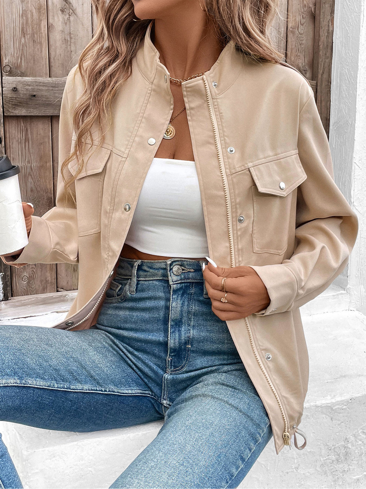 Person wearing a beige jacket, white top, and blue jeans, holding a coffee cup.