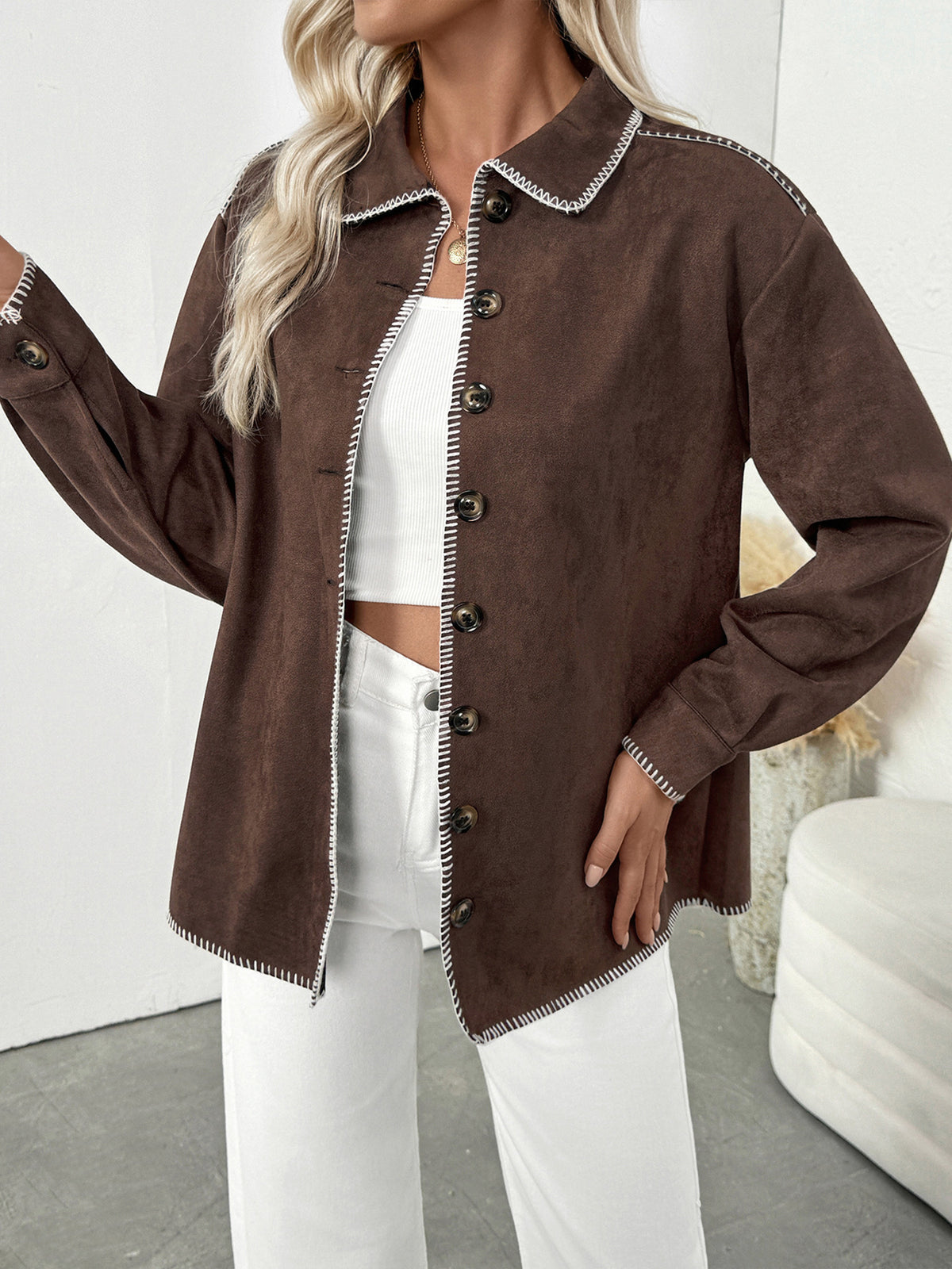 A woman wearing a brown faux-suede button-up shirt jacket with white trim and dark buttons, styled over a white crop top and white pants.