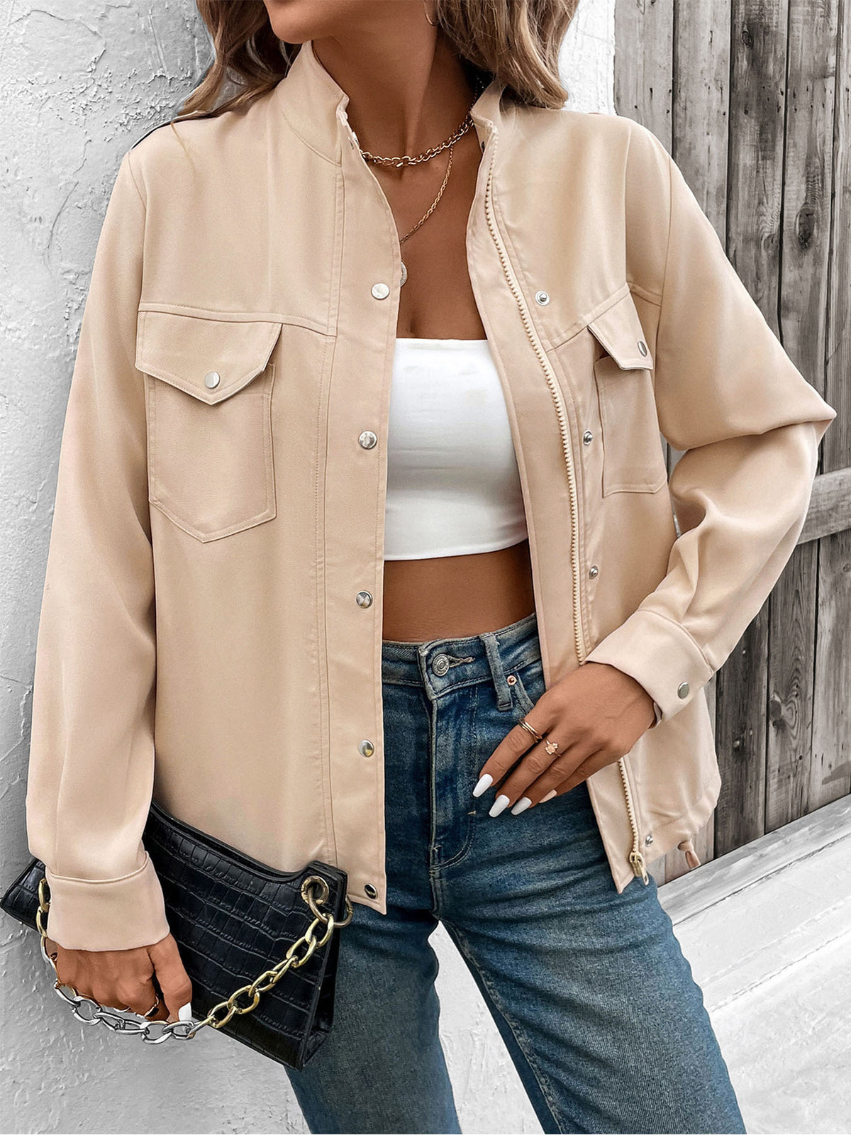 Person wearing a beige jacket, white crop top, and blue jeans against a wooden fence background.
