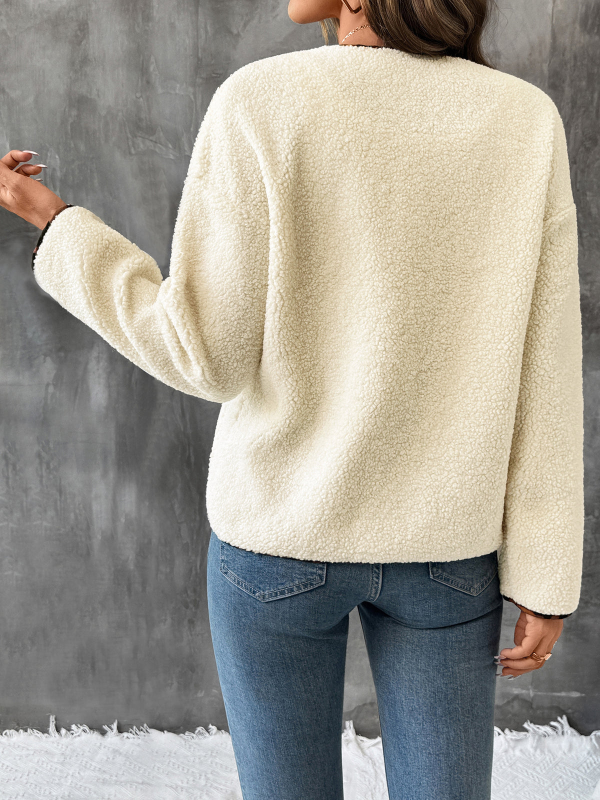 Person wearing a cream-colored sweater and blue jeans against a gray wall.
