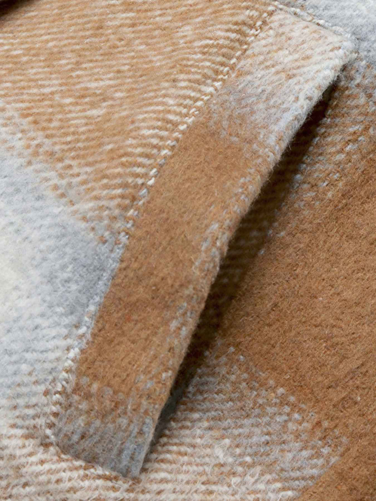 Close-up of a soft woven textile with a plaid pattern in warm tan, beige, cream, and light gray, showing the fabric texture and folded edges.