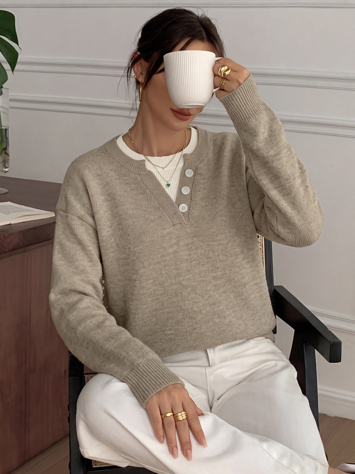 A woman sitting in a chair wearing a light beige v-neck knit sweater over a white top and white pants, holding a white mug to her face in an indoor setting.