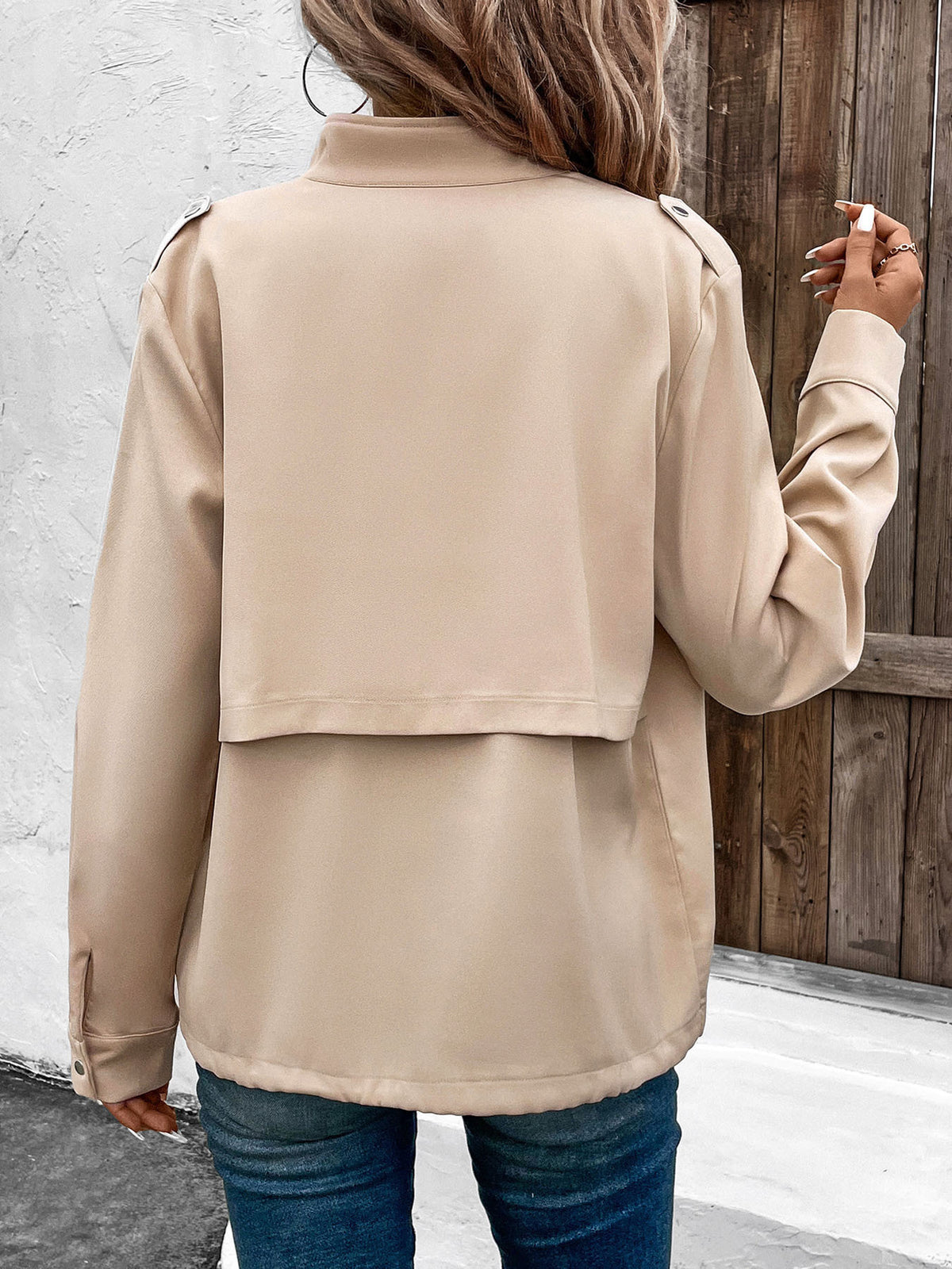 A woman viewed from behind wearing a beige long-sleeve cropped jacket with shoulder epaulets, paired with blue jeans, standing in front of a white wall and wooden door.