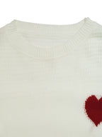 Close-up of a cream-colored knit sweater with a textured waffle pattern and a small red heart knit on the lower right chest, set against a plain white background.