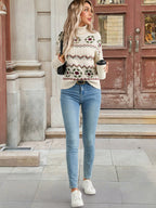 Woman wearing a patterned sweater and jeans, holding a coffee cup, standing in front of a building entrance.