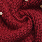 A close-up of a chunky knit fabric in deep burgundy red, styled in a swirl pattern with small white pearl accents.