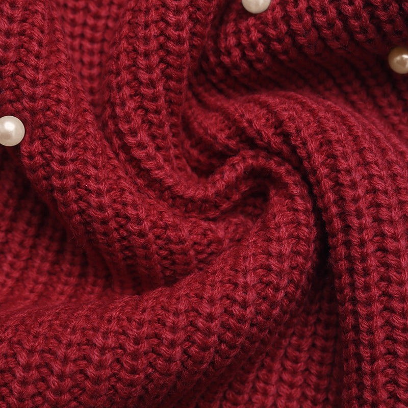 A close-up of a chunky knit fabric in deep burgundy red, styled in a swirl pattern with small white pearl accents.