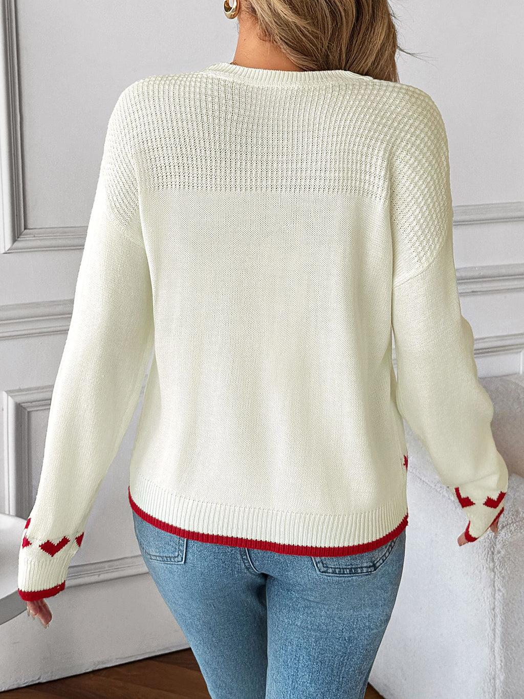 Person wearing a white sweater with red accents and blue jeans against a gray wall.