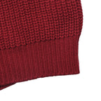 Close-up detail of a chunky knit garment in a deep burgundy red, showing the ribbed cuff against a plain white background.