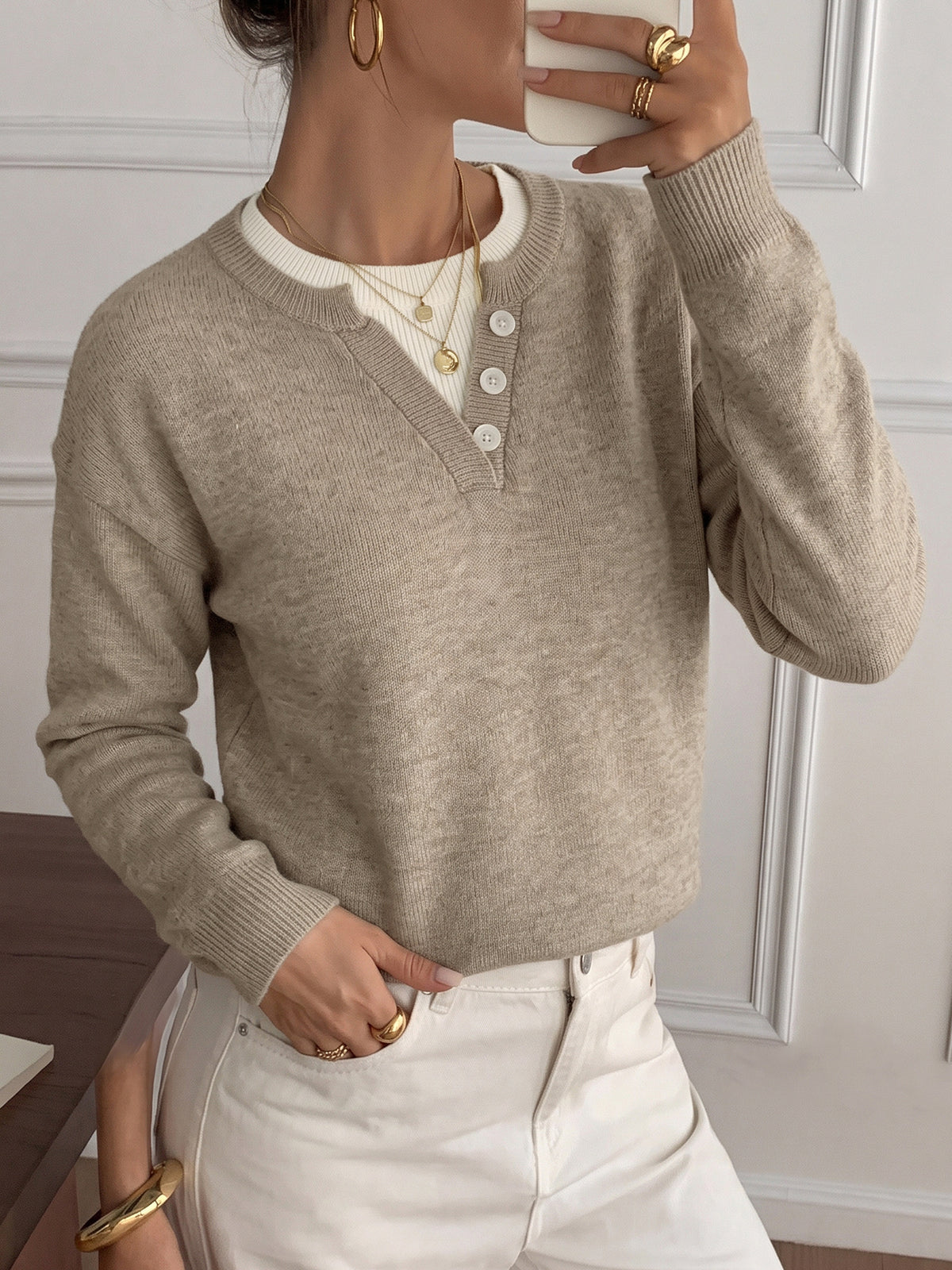 A woman wearing a light taupe knit henley sweater over a white ribbed top and white pants, holding a white mug in a home interior setting.
