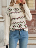 A woman wearing a cream-colored knit sweater with a colorful Nordic-style geometric pattern in green, burgundy, and brown, paired with blue jeans.