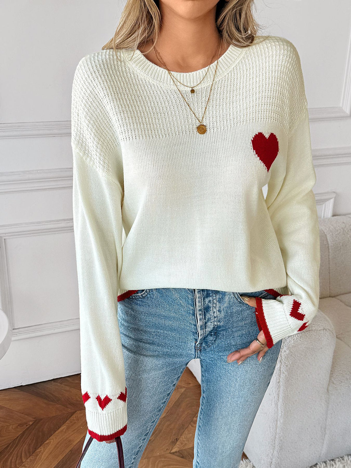 A woman wearing a cream-colored knit sweater with red heart details on the chest and cuffs, paired with light-wash blue jeans.