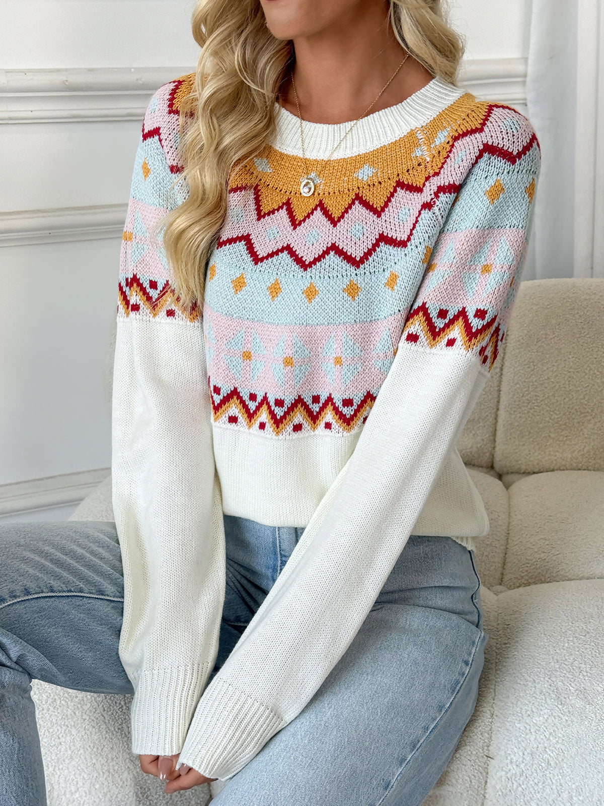 A woman wearing a long-sleeve knit sweater with a multicolor Fair Isle pattern on the chest and sleeves, paired with light blue jeans, sitting on a beige couch.