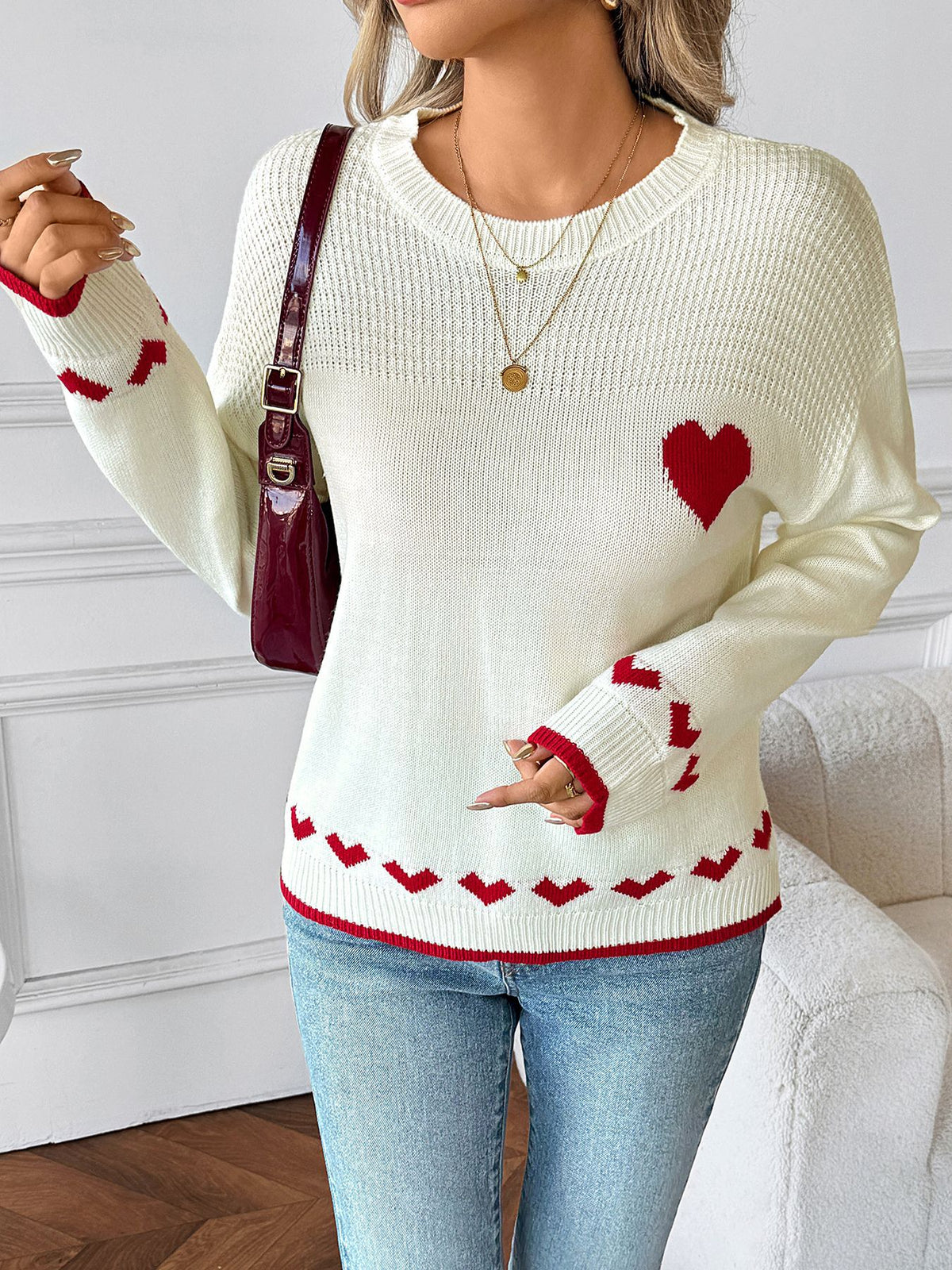 Woman wearing a white sweater with red heart designs, holding a maroon handbag indoors.