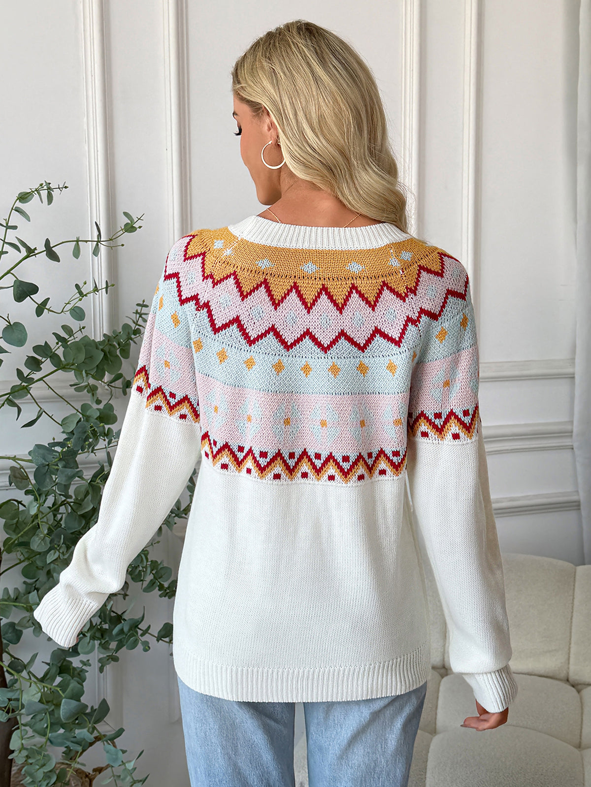 Woman wearing a white sweater with colorful pattern, standing indoors next to a plant.