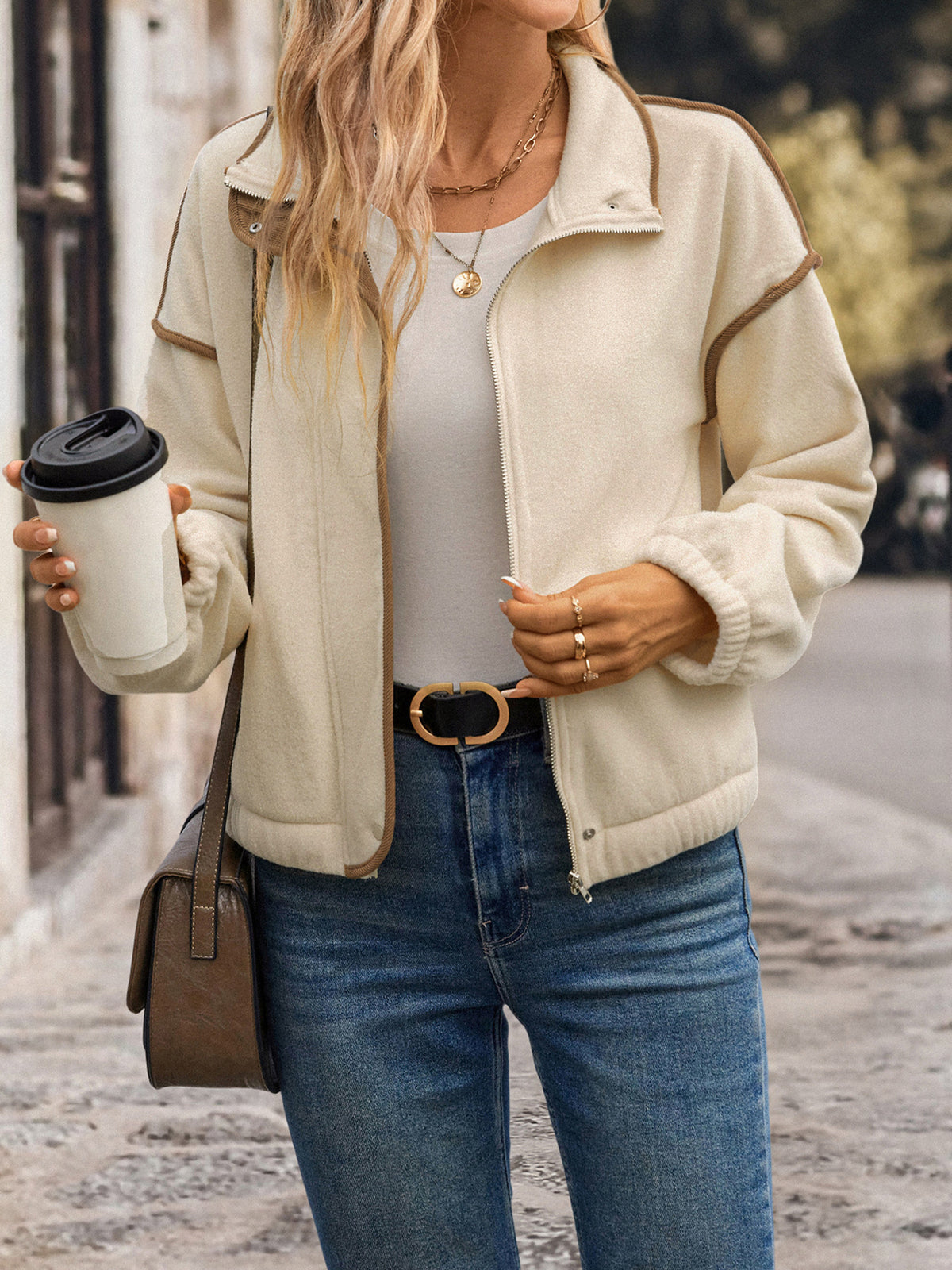 Person wearing a beige jacket, white shirt, and blue jeans, holding a coffee cup.