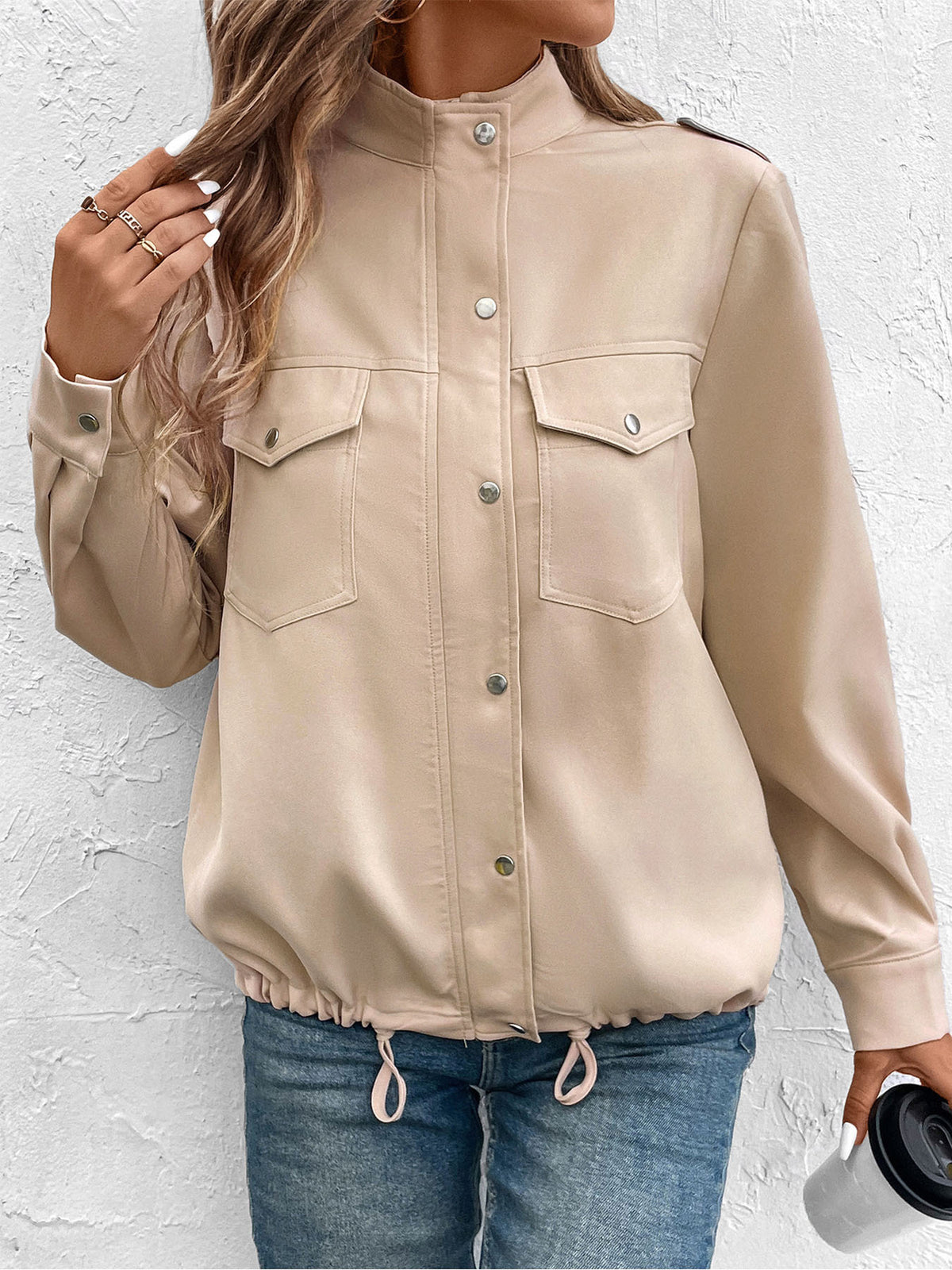 A woman wearing a beige long-sleeve button-front jacket with flap chest pockets and a drawstring hem, paired with blue jeans, standing against a textured white wall.